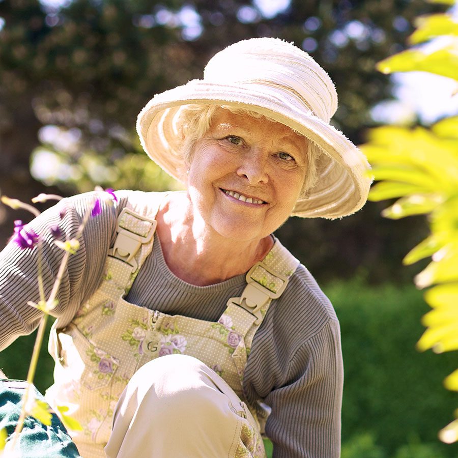 An older woman wearing a sun hat and overalls smiles while gardening outdoors, surrounded by greenery and flowers in bright sunlight.