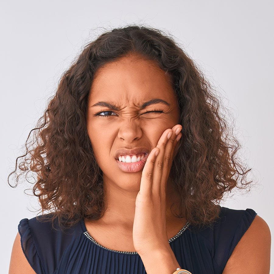 A young woman with curly hair touches her cheek and winces in pain, suggesting she has a toothache or facial discomfort. She is wearing a navy blue top and standing against a plain light background.