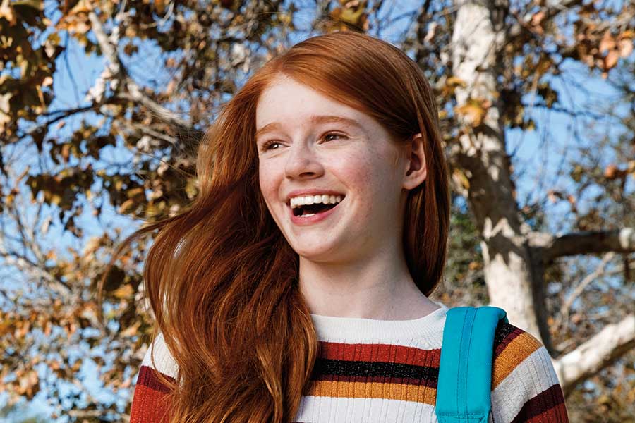 A woman sitting in a dental chair smiles while holding a clear orthodontic aligner. She wears a dental bib and appears to be at a dental clinic.