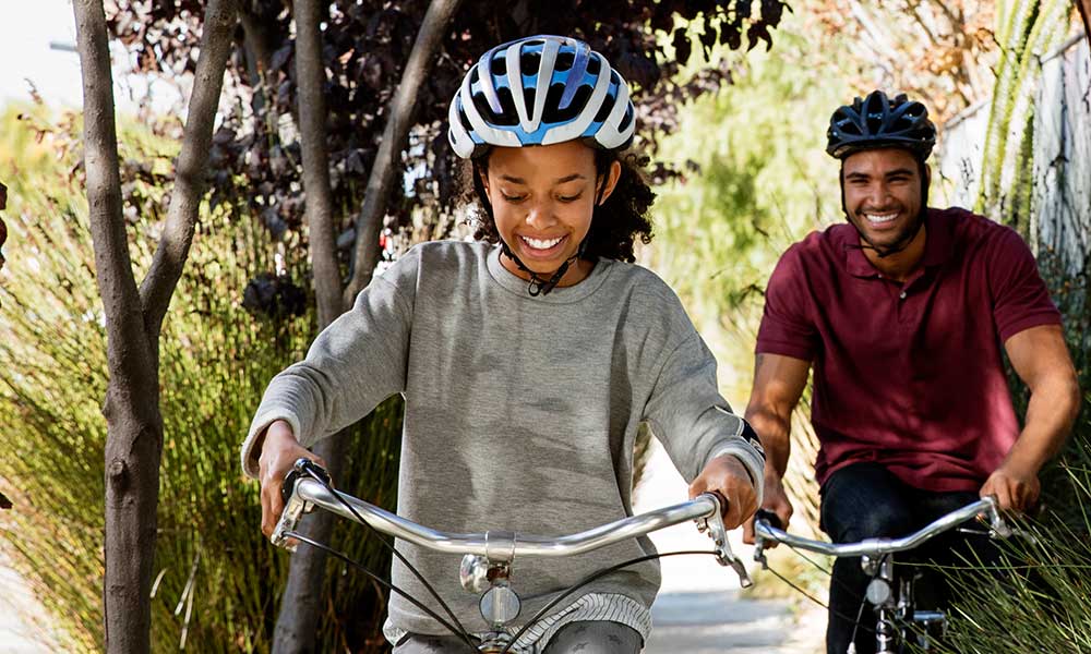 A man and a woman cycling together on a tree-lined path, enjoying a sunny day outdoors.