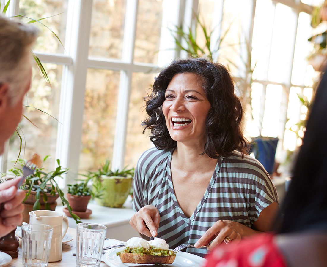 A woman with dark, curly hair smiles and laughs while sitting at a table and eating brunch with others in a bright room filled with potted plants and natural light.