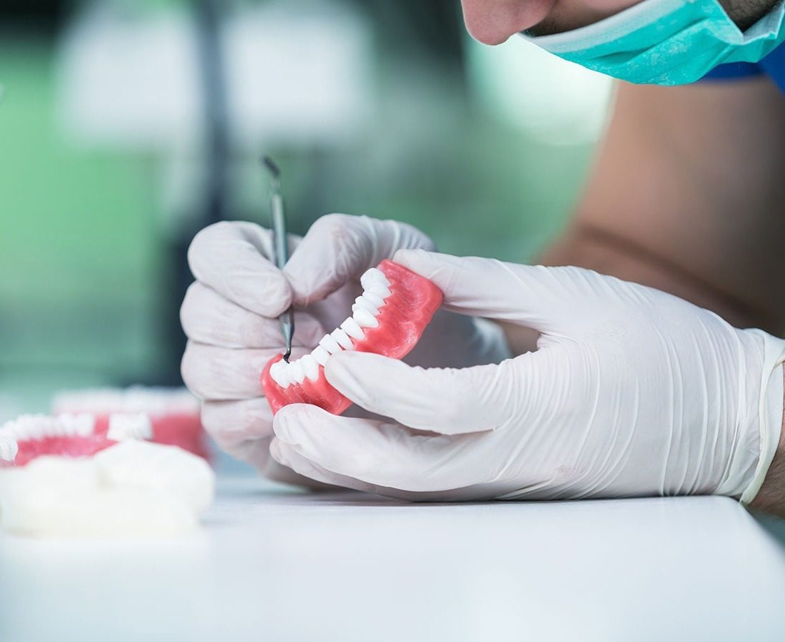 A person wearing gloves and a face mask uses a dental tool to work on a set of artificial dentures, holding them carefully on a white table.
