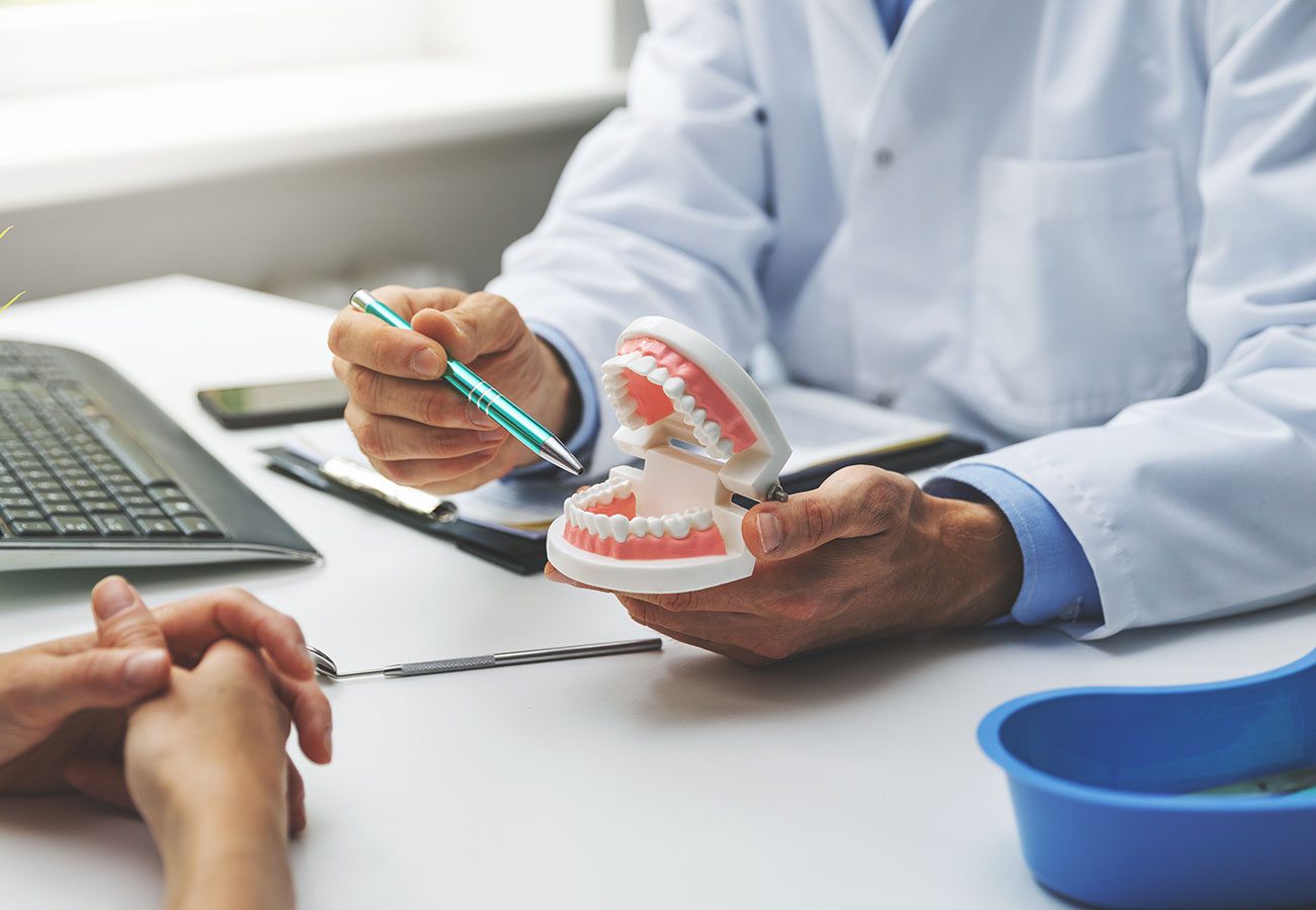A dentist in a white coat holds a dental model and points at it with a pen while explaining something to a patient across the desk. Dental tools and a computer are visible on the table.