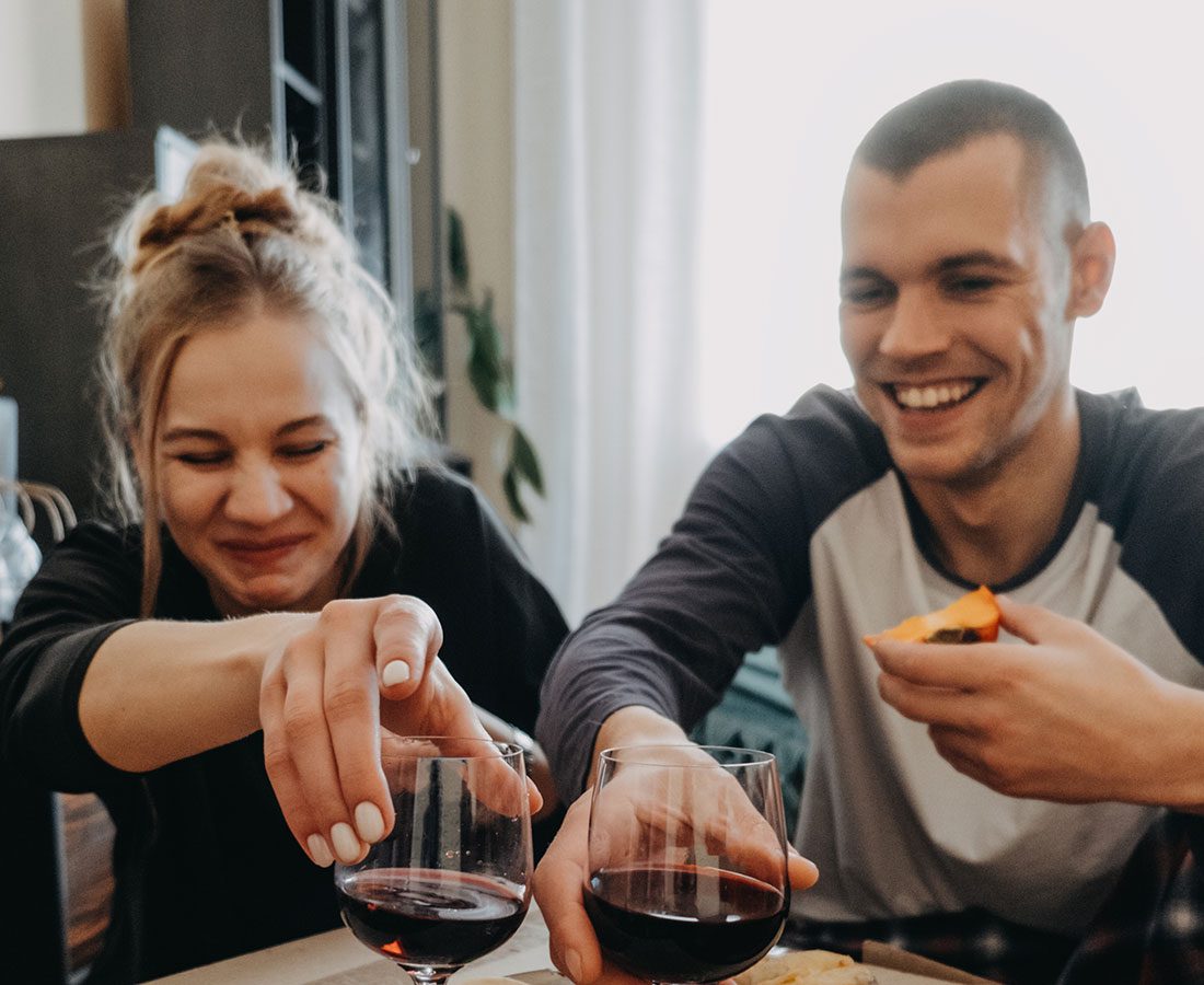Two people sit close together, smiling and laughing while holding glasses of red wine. One person is also holding a slice of pizza. The atmosphere appears casual and cheerful.