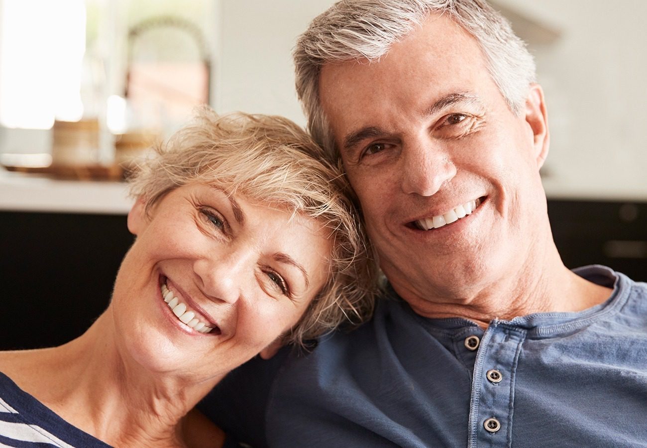 A smiling older couple with gray hair sits closely together indoors, looking at the camera. The woman leans her head on the man’s shoulder. They both wear casual shirts and appear happy and relaxed.