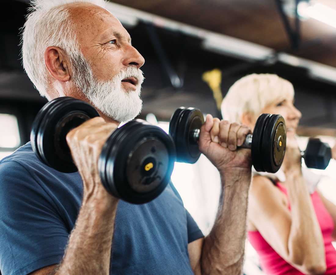 An older man with a white beard lifts dumbbells at a gym, smiling and wearing a blue shirt. A woman in the background also lifts weights, both appearing focused and active.