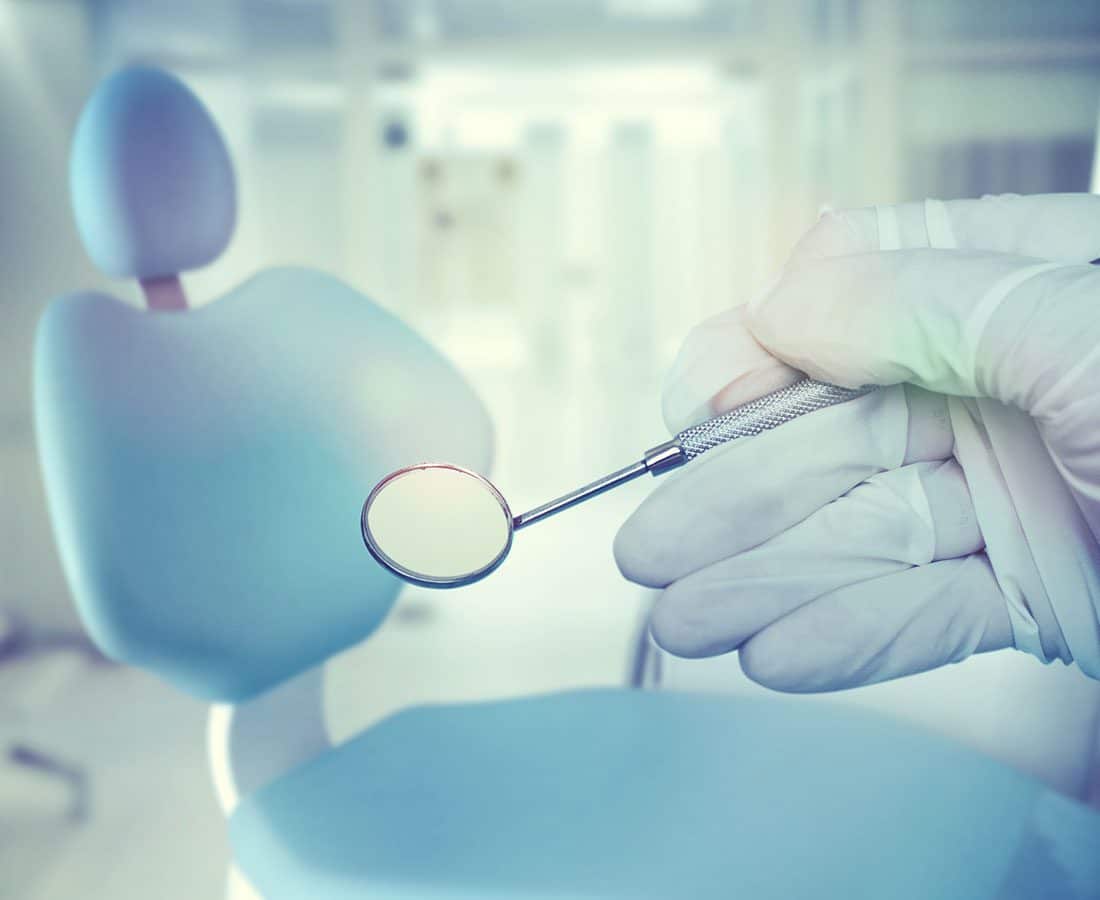 A gloved hand holds a dental mirror in the foreground, with a blurred dental chair and clinical equipment visible in the background.