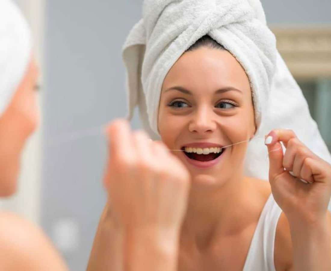 A smiling person with a towel wrapped around their head is looking in the mirror and using dental floss, appearing to clean their teeth in a bathroom.