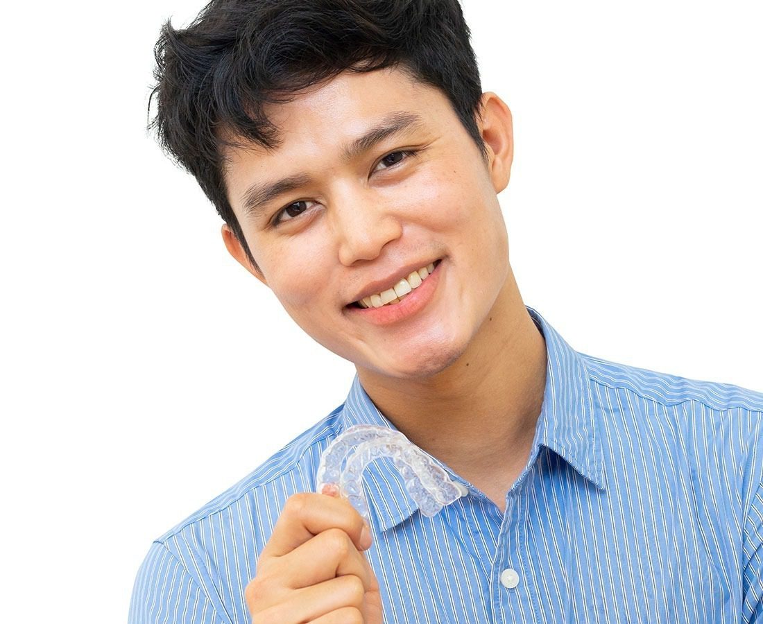 A smiling person with short dark hair, wearing a blue striped shirt, holds up a clear dental aligner against a white background.