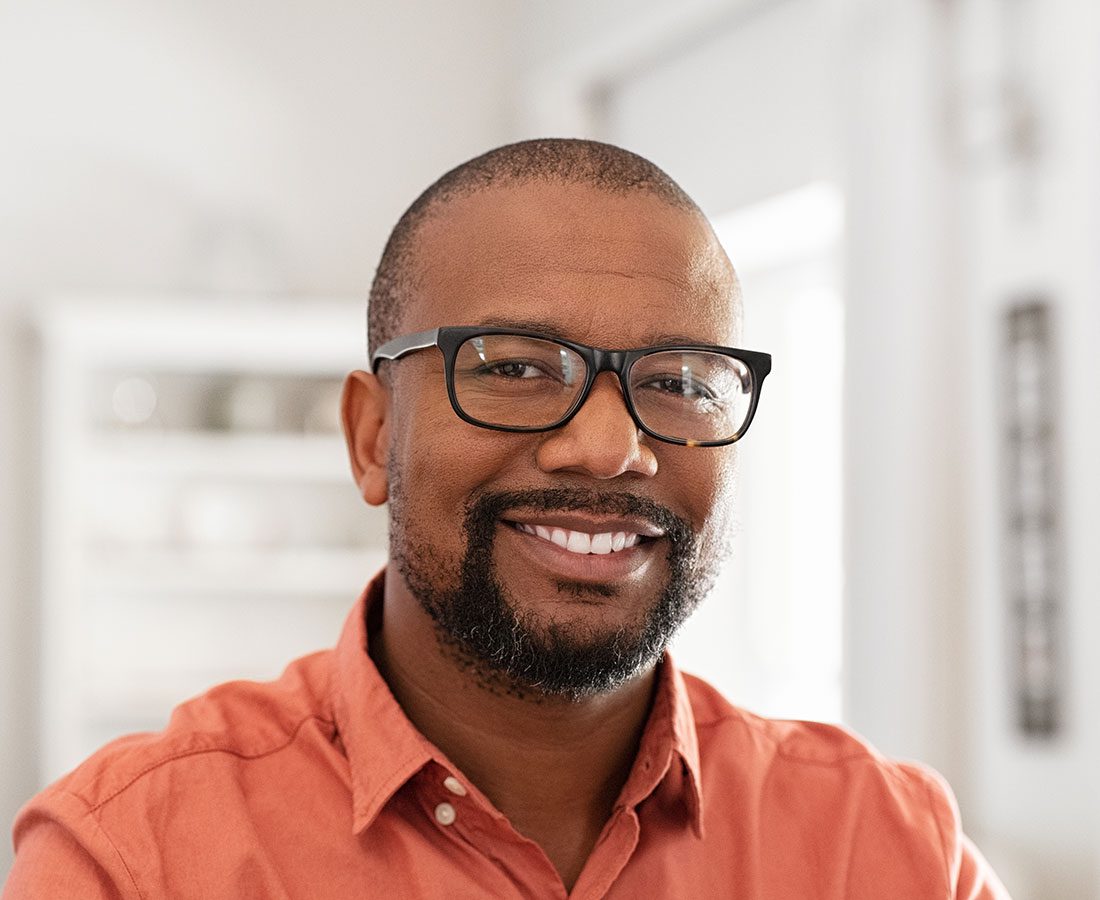 A smiling man with short hair, a neatly trimmed beard, and glasses, wearing an orange shirt, stands indoors in a softly lit room with a blurred background.