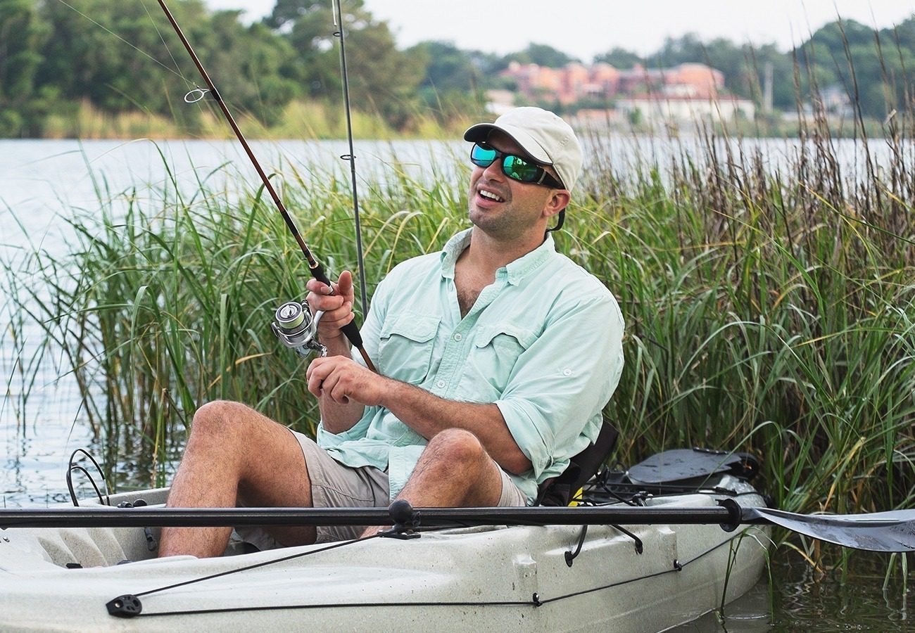A man wearing sunglasses and a light green shirt sits in a kayak, fishing and smiling, surrounded by tall grass on a calm lake with trees and buildings in the background.