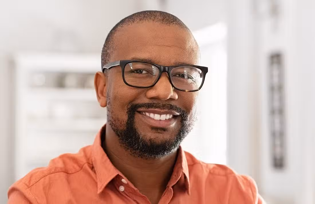 A smiling man with short hair, a trimmed beard, and glasses, wearing an orange shirt, stands indoors in a well-lit room with a blurred background.