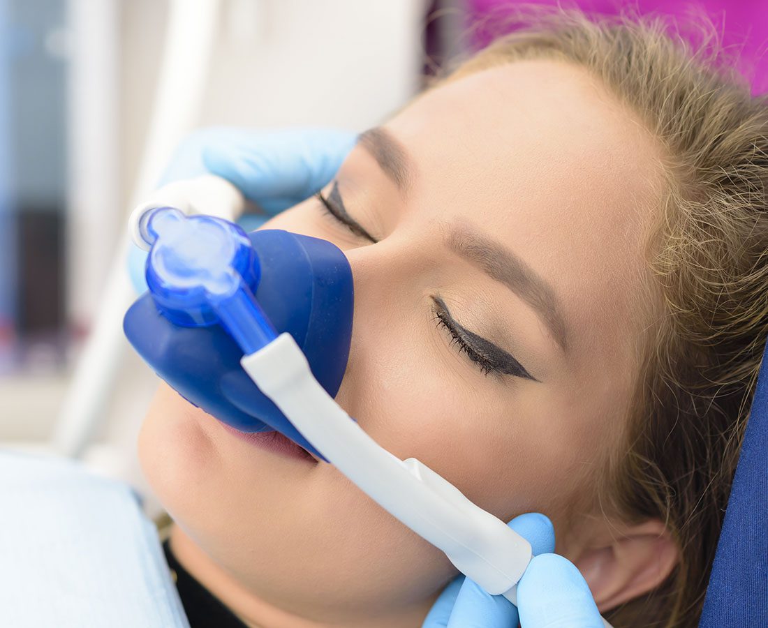 A woman with closed eyes is wearing a blue and white nasal mask for sedation, likely at a dental office. She is reclined in a chair with gloved hands adjusting the mask on her face.