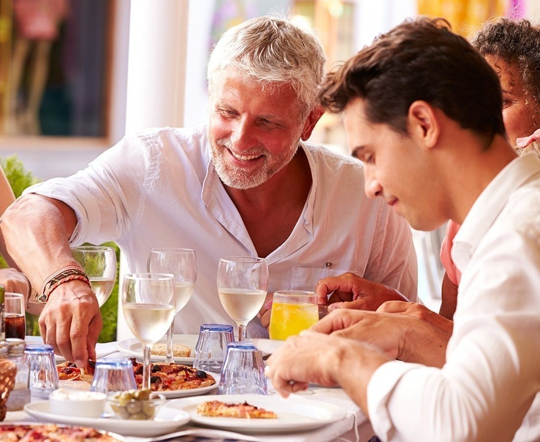 Two men are enjoying a meal together at an outdoor restaurant, smiling and sharing food. There are plates of pizza, glasses of white wine, and orange juice on the table. Other people are partially visible in the background.