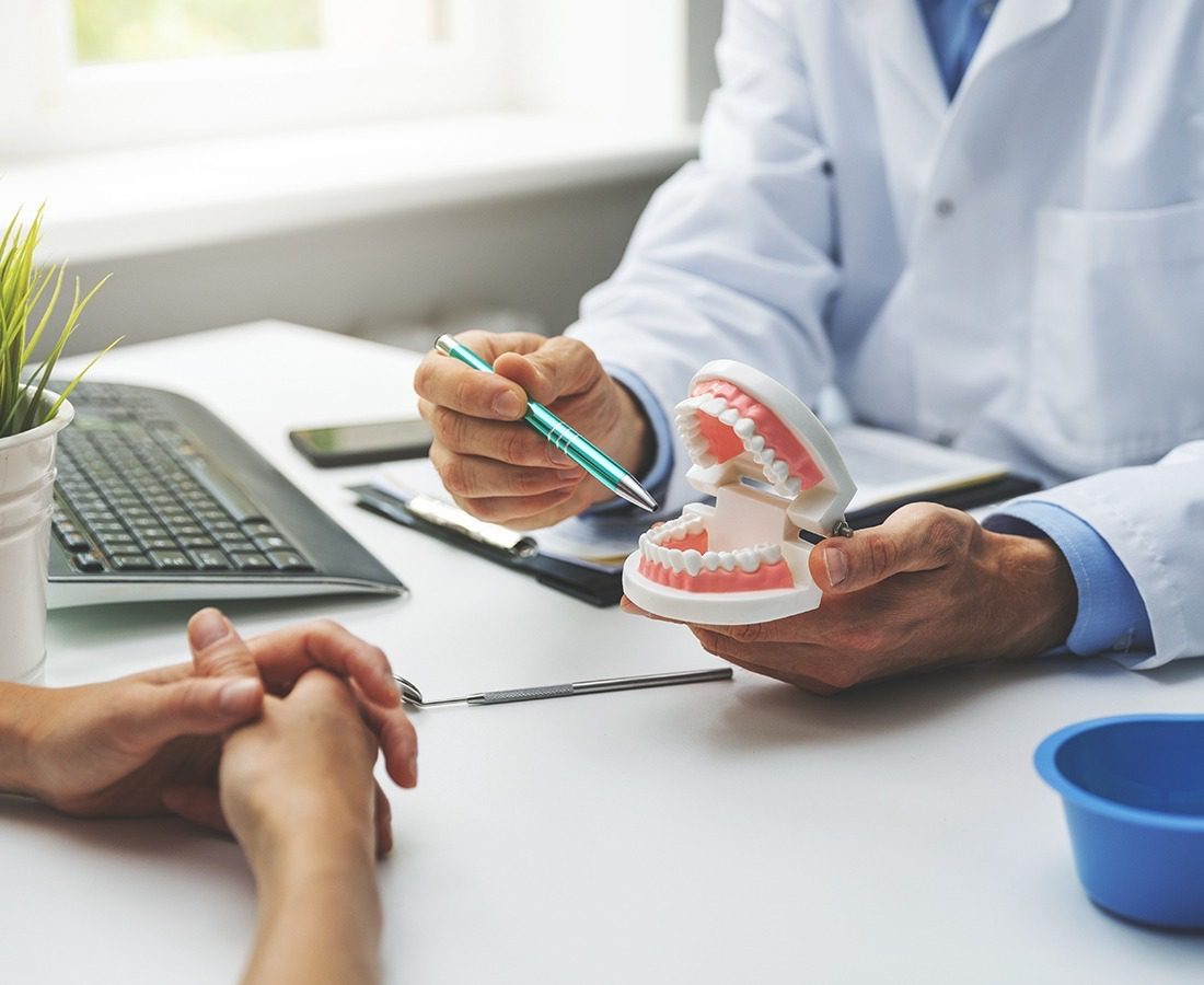 A dentist in a white coat explains dental care using a model of human teeth, holding a pen and gesturing, while a patient listens across the desk in a bright, modern office.