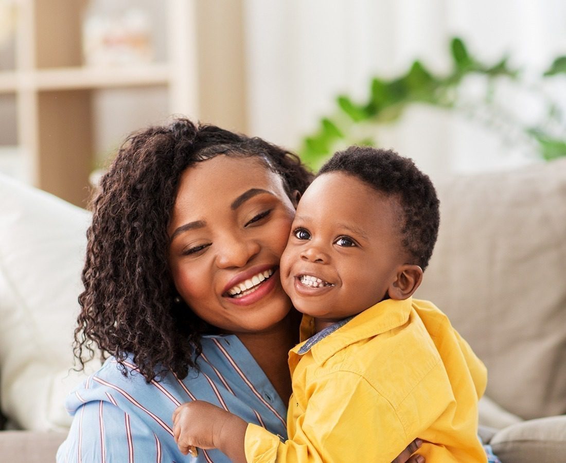 A smiling woman hugs a cheerful young boy in a bright, cozy living room. The woman wears a blue striped shirt, and the boy wears a yellow shirt. Green plants and shelves are blurred in the background.