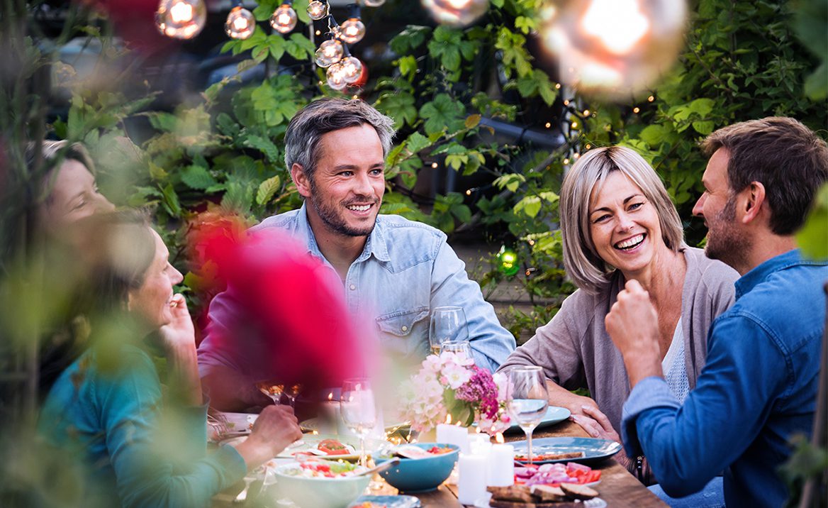 Four people sit at an outdoor table, laughing and enjoying food together amid greenery and string lights, creating a warm, festive atmosphere.