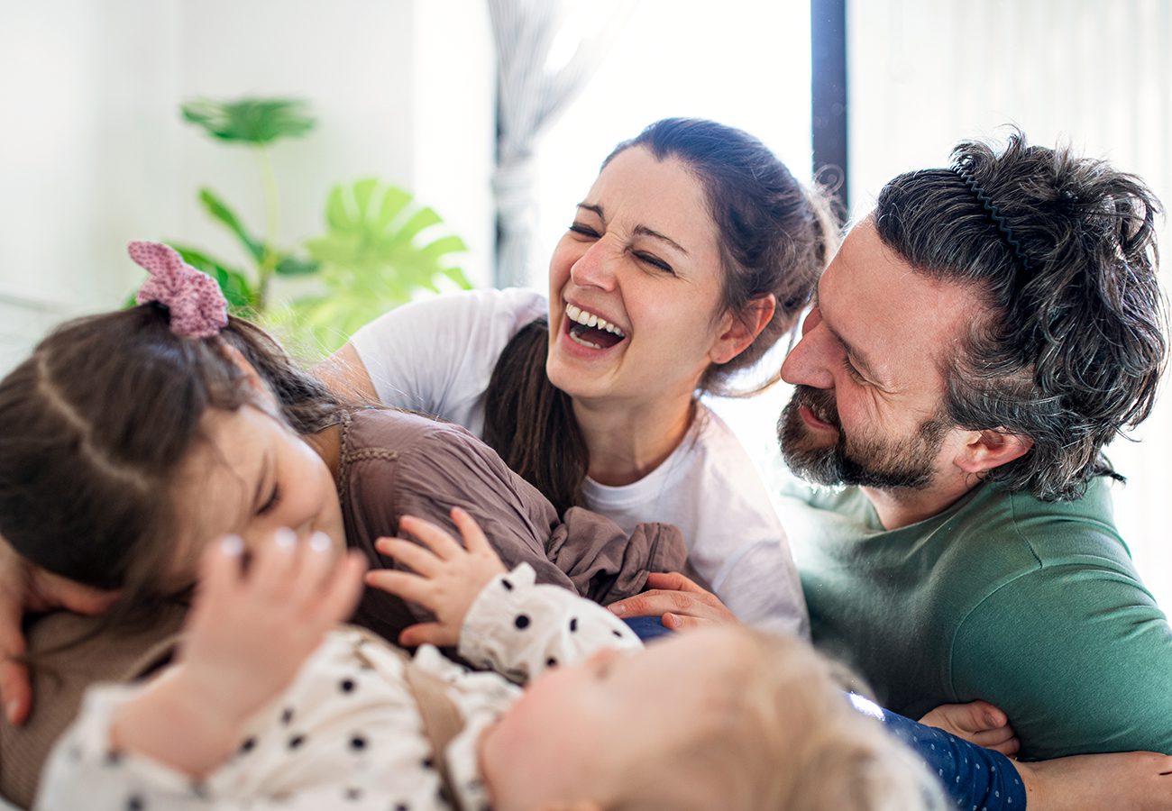 A joyful family of four, including two adults and two children, laugh and hug together indoors. The parents smile warmly while the children play and giggle, creating a lively and happy moment.