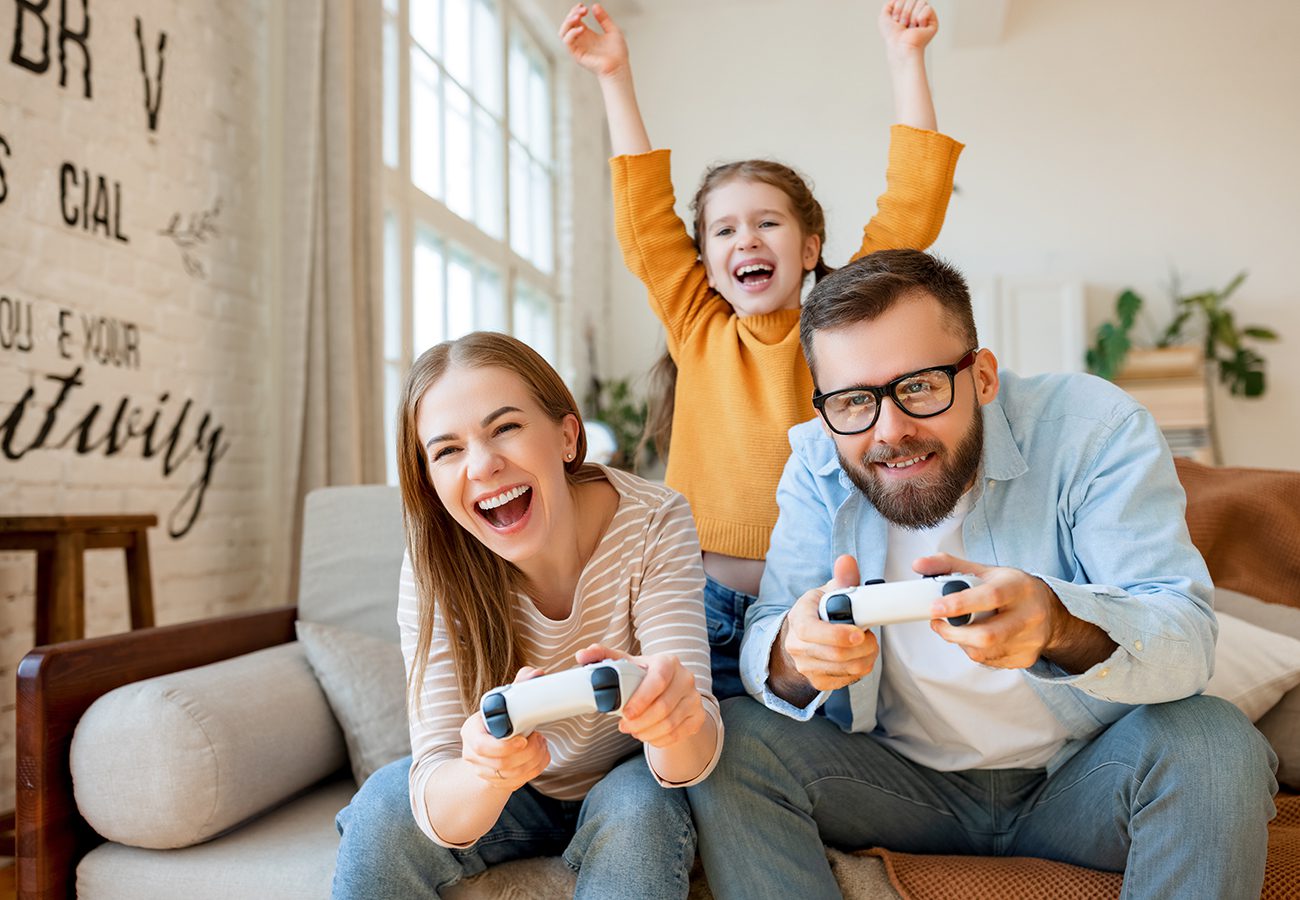 A smiling man and woman play video games with controllers on a couch while a young girl behind them cheers with her arms raised, all appearing happy and excited in a bright living room.