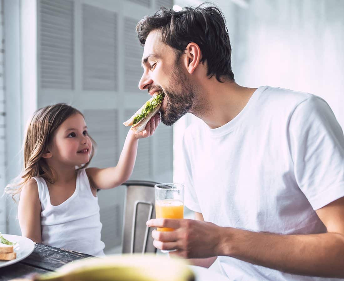 A young girl in a white tank top feeds a sandwich to a smiling man holding a glass of orange juice. They sit together at a table, enjoying a meal in a bright, airy room.