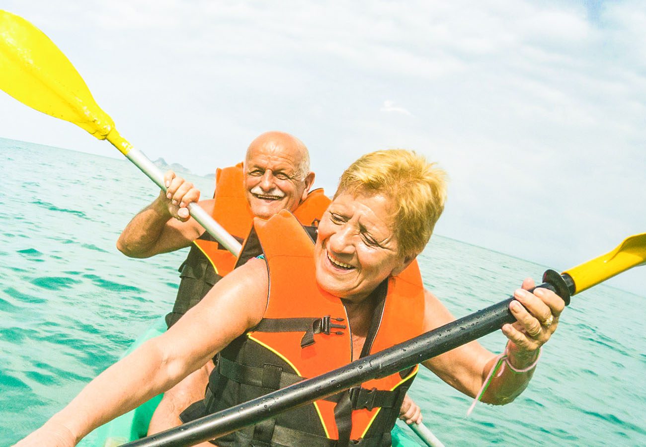 An older man and woman, both wearing orange life jackets, paddle a kayak together on clear blue water, smiling and enjoying a sunny day outdoors.