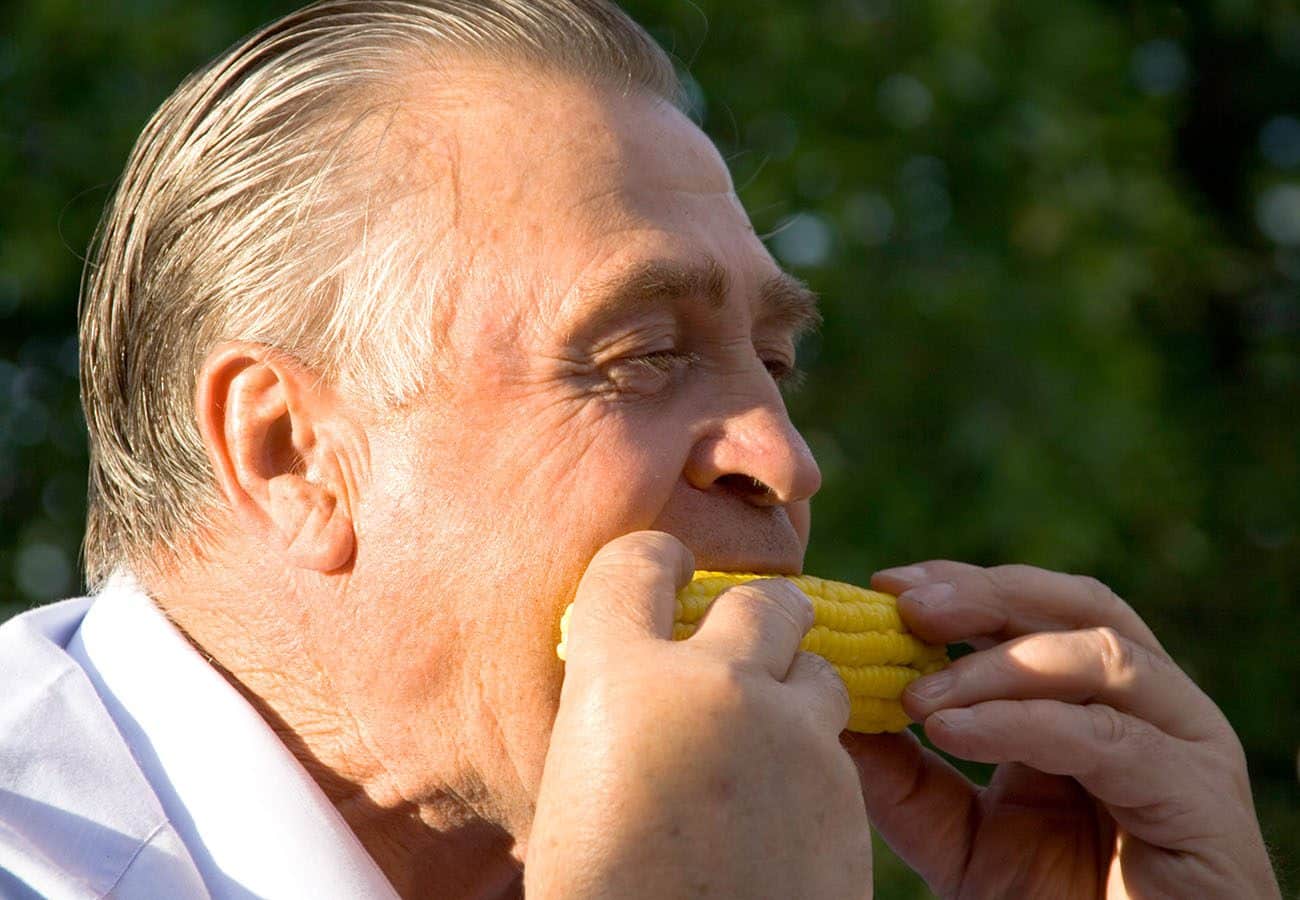 An older man with gray hair and a white shirt eats a corn on the cob outside in bright sunlight, with green foliage blurred in the background.