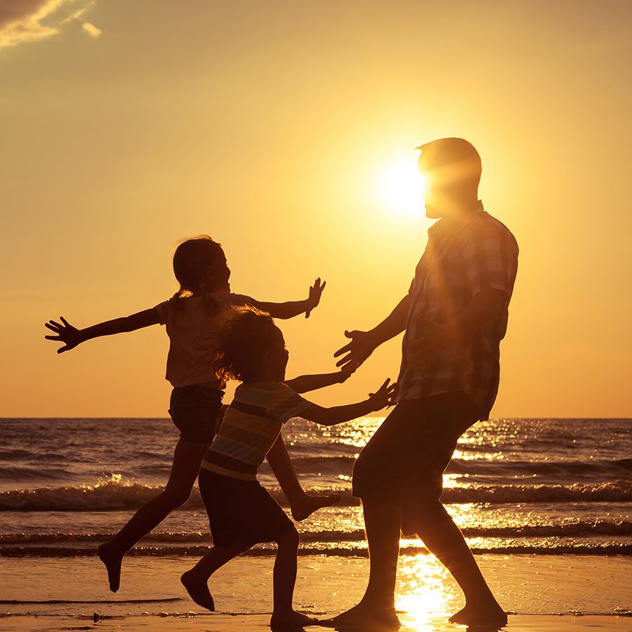 A silhouette of an adult and two children playing together on a beach at sunset, with the sun low in the sky and the ocean in the background.