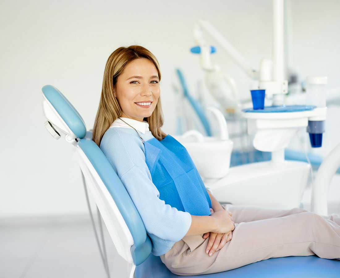 A woman sits in a dentists chair, smiling and wearing a blue dental bib. Dental equipment and a cup are visible in the bright, clean clinic background.